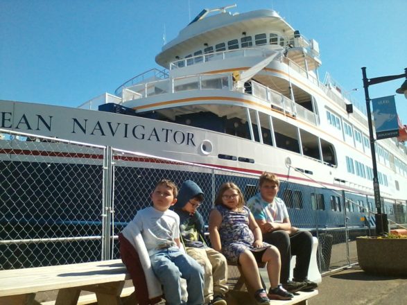 Oliver, Justin, Olivia, and Reggie sitting on a bench in front of the cruise ship at the Houghton canal.
