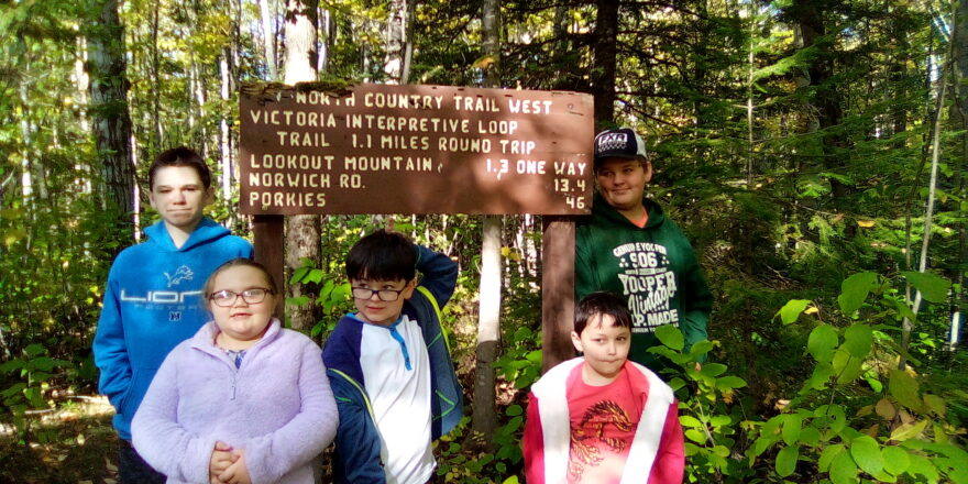 Reggie, Olivia, Justin, Oliver, and Jacob standing around the sign for the North Country Trail by the sauna.