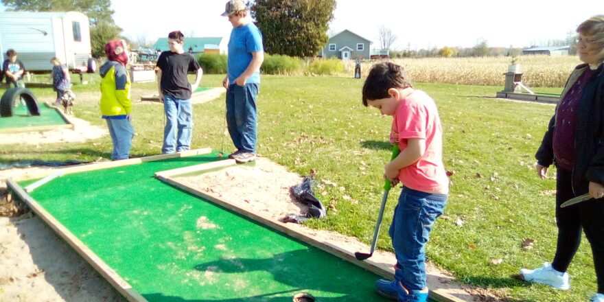 Oliver is taking his turn putting with Ms. J. giving him pointers, while Avery, Jacob, and Reggie wait by the hole on the putting green.
