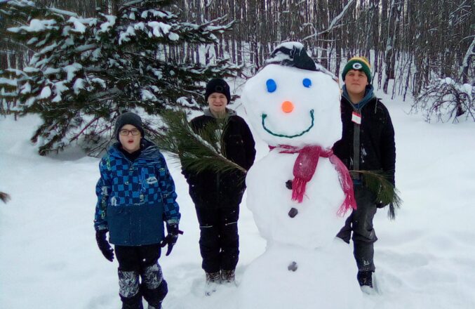 The 3 older kids standing with Snowy the snowman in his winter hat, cups for blue eyes, paint can top orange nose, green wire smiley mouth, purple scarf, pine branches for arms, and black buttons of foam.