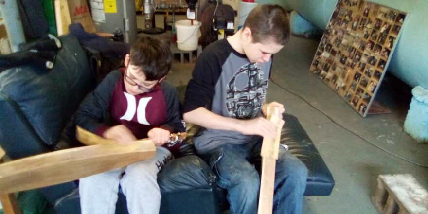 Avery and Jacob sitting side by side and sanding their freshly made dulcimers by hand.