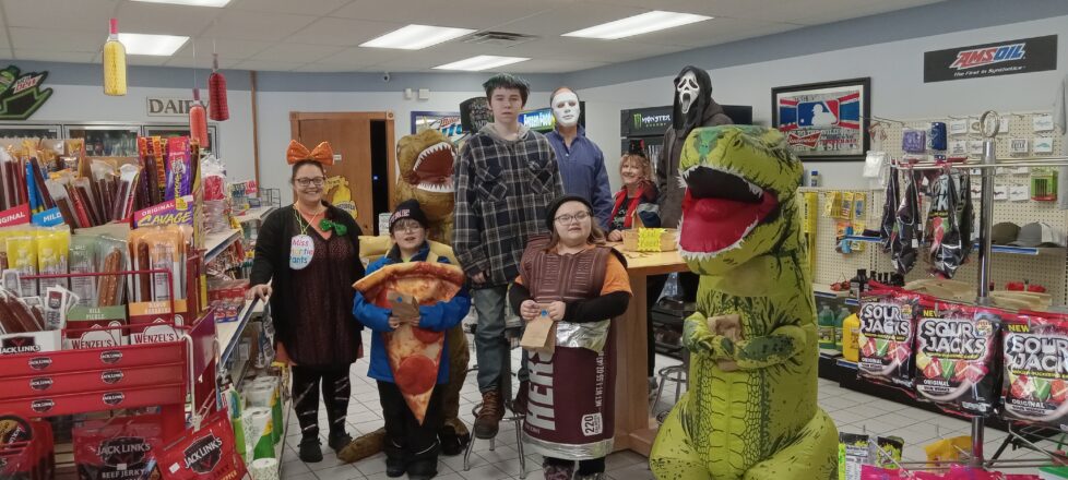All the kids in their Halloween costumes. Standing at the table in 4 seasons gas station are a slice of pepperoni pizza, an Hershey's bar, two inflatable dinosaurs, a ghost face, and a Michael Meyers costume for our bus driver.