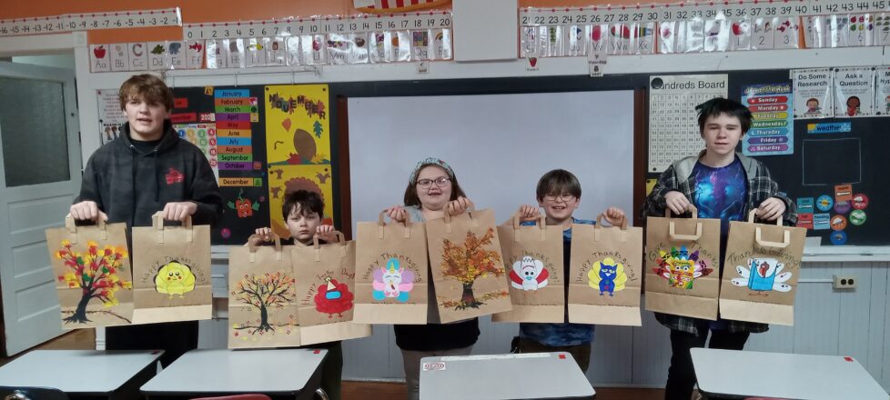 Kids lined up in front of the room to pose for a picture with their decorated thanksgiving bags. Each of the kids are holding a different bag in each hand that they had decorated. Some have fall colored leaves on the trees and others have a picture of a turkey in disguise.