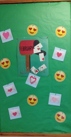 Board in entrance for month of February. Has mailbox with love letters falling out of it. Smiley faces with heart eyes and letters with hearts on them scattered around the board.