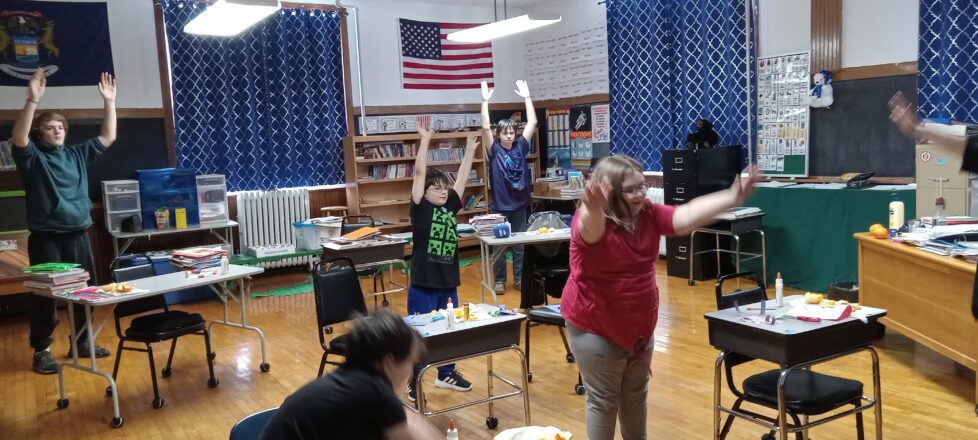 Kids are standing by their desks and raising their arms up in the air to do exercises.