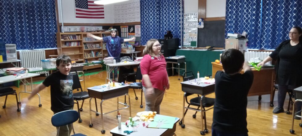 Kids are standing by their desks and doing jumping jacks.