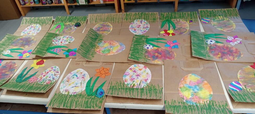 Easter bags displayed on table that were decorated by kids. Brown paper bags have grass painted on the bottoms with a watercolor splatter paint design and next to them is a colored flower.