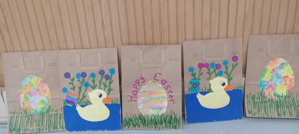 Brown bags showing Easter decorations. Some of the bags have a big brightly water colored egg in the middle sitting on grass. The others have a white duck with yellow beak sitting in a little pond with flowers growing on the edge behind the duck.